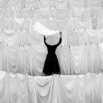 Black and white photo of a person wearing a conical hat standing among rows of hanging white sheets on clotheslines, lifting a cloth overhead, creating a striking pattern of repeating fabric textures and minimalist contrast.